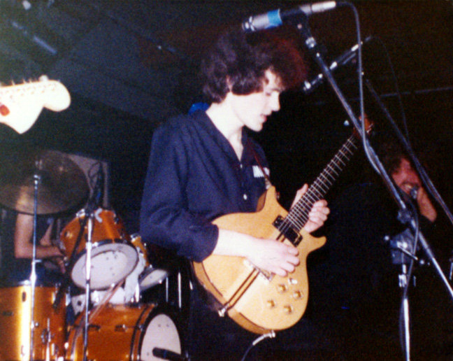 A young guitarist with long curly hair playing a natural-finish electric guitar on a dark stage, drum kit behind him.